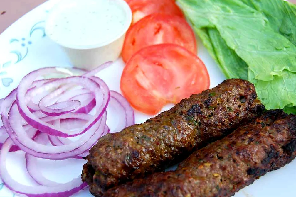 Two seekh kababs on a plate with sliced tomato, red onion rings, lettuce and a cup of raita